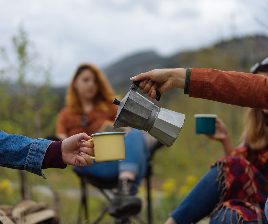 Un gruppo di amici all'aperto condivide del caffè, in primo piano una moka che viene versata in una tazza , sullo sfondo, una ragazza dai capelli rossi e montagne.